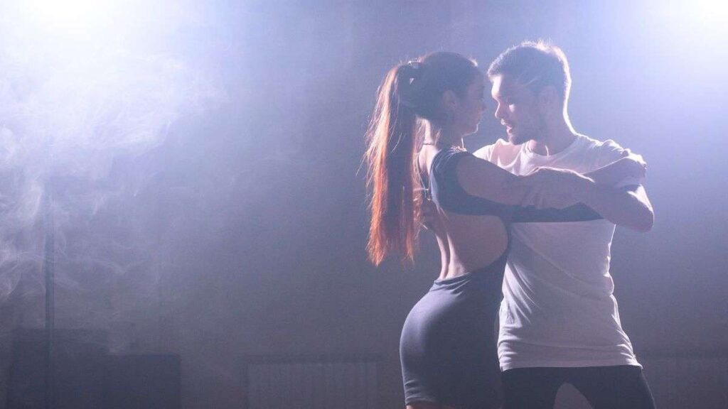 Pár tančí bachatu na taneční lekci, světlo na něho svítí a kolem něj se vznáší kouř.